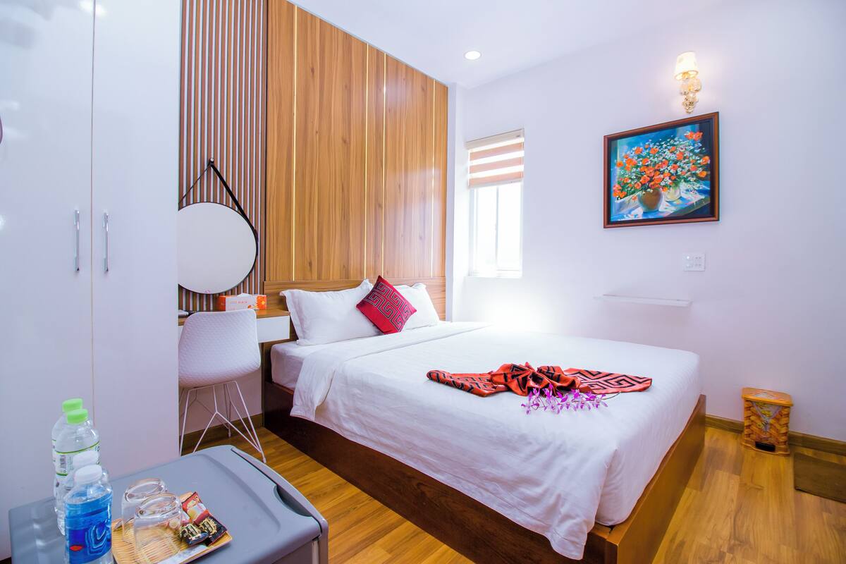 Ein Hotelzimmer mit einer Wandpaneele aus Holz, einem Bett mit weißer Bettwäsche, einem Blumenbild an der Wand und einem kleinen Tisch mit einer Wasserflasche und Snacks.