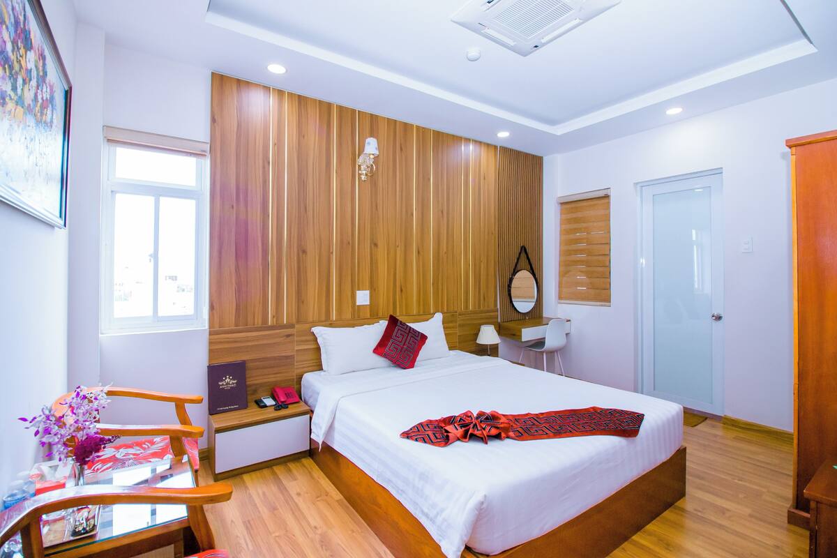 Ein modernes Hotelzimmer mit einem Holzkopfteil, einem großen Bett mit weißer Bettwäsche, einem Schreibtisch mit Stuhl und einem Fenster, das natürliches Licht hereinlässt.