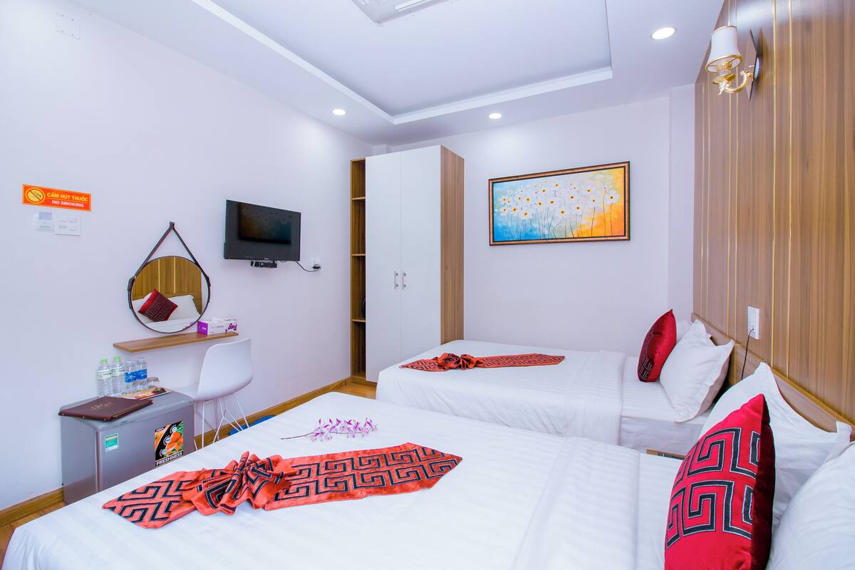 Ein Hotelzimmer mit zwei Betten, einem Fernseher, einem Schreibtisch mit Stuhl, einer kleinen Kühlbox und einem Bild an der Wand.