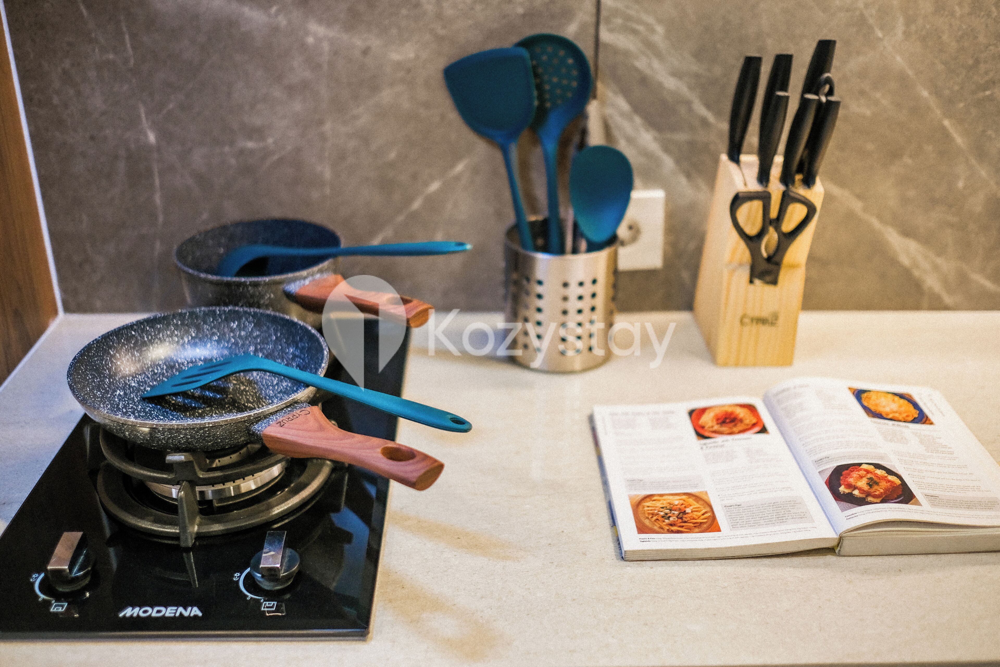 Una cocina con estufa, sartén, un libro y utensilios de cocina.