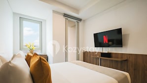 A modern hotel room with a bed, a TV mounted on the wall, a window with a view, and a wooden shelf. - Staya Antasari by Kozystay - Cilandak (Jakarta)