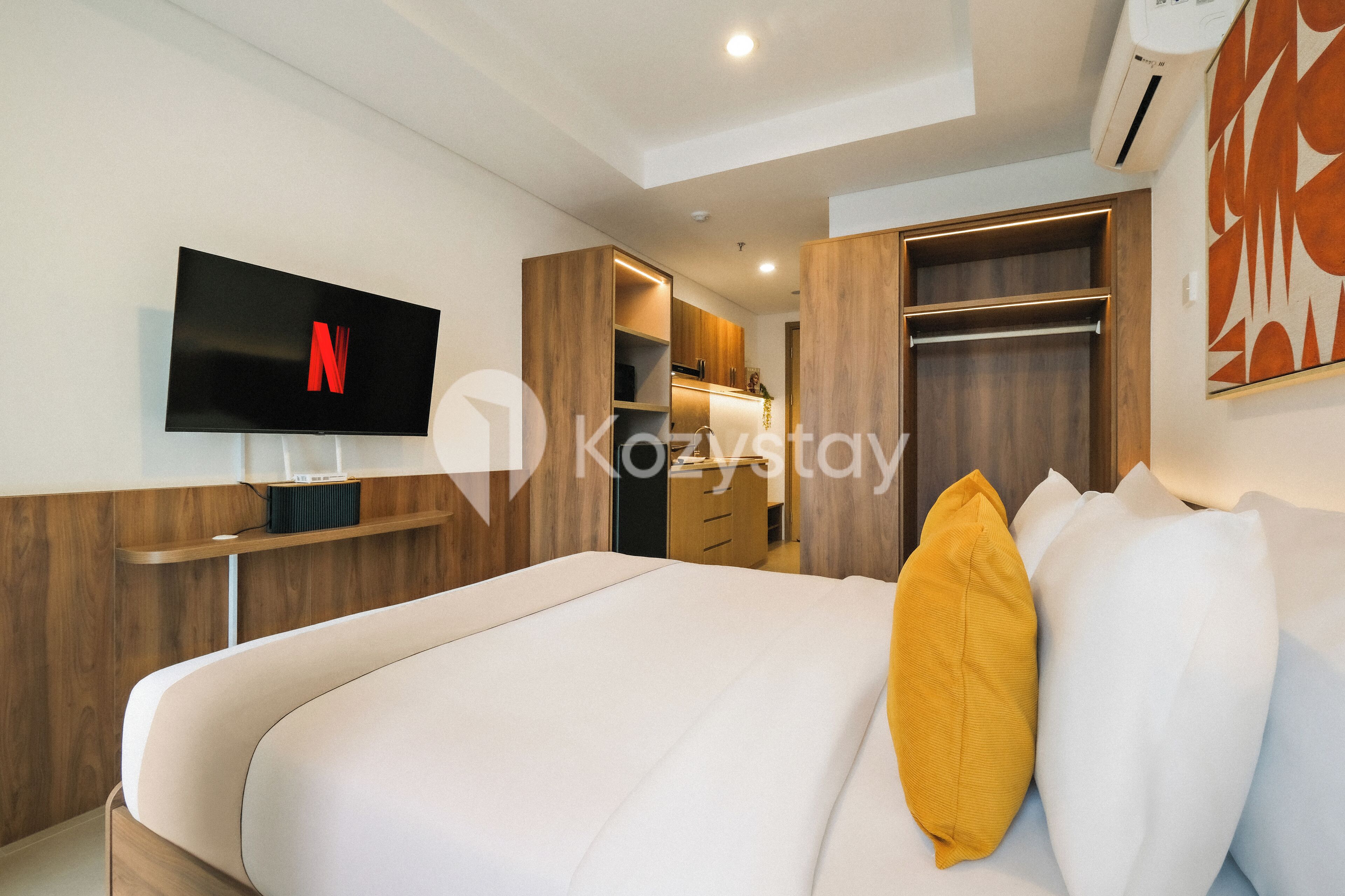 A modern hotel room with a flat-screen TV mounted on the wall, a wooden wardrobe, a built-in closet, and a bed with white and yellow pillows.