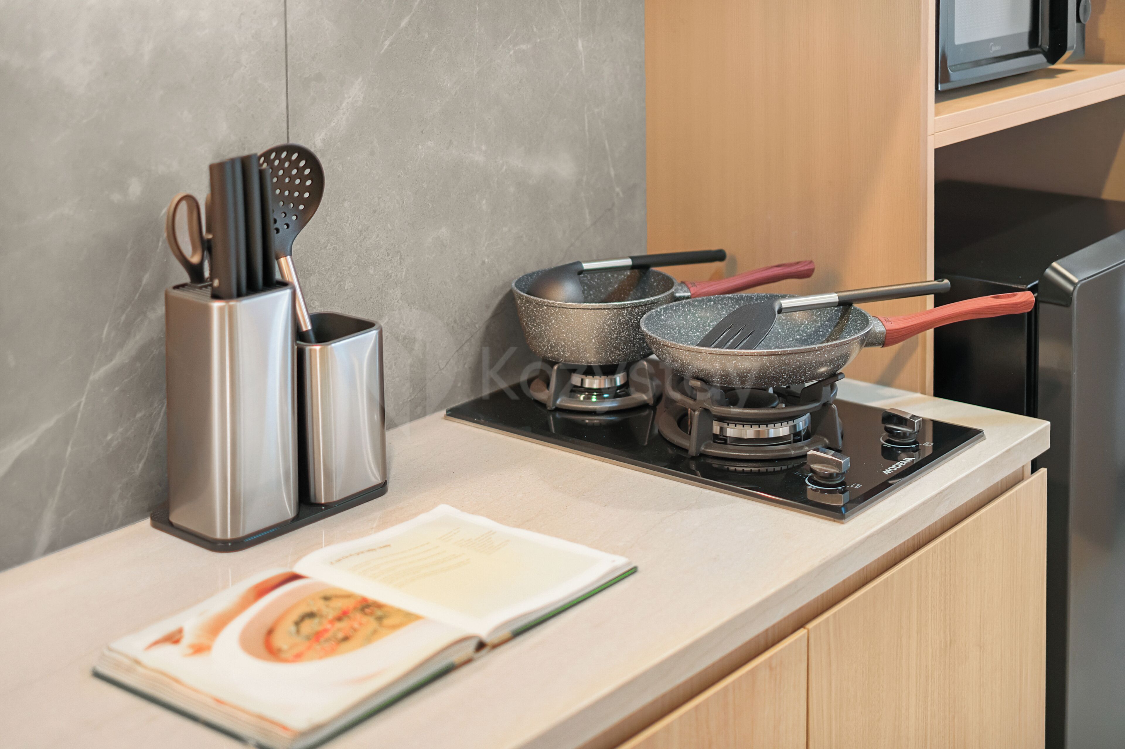 A kitchen counter with a stove, two frying pans, a spatula, and a cookbook.