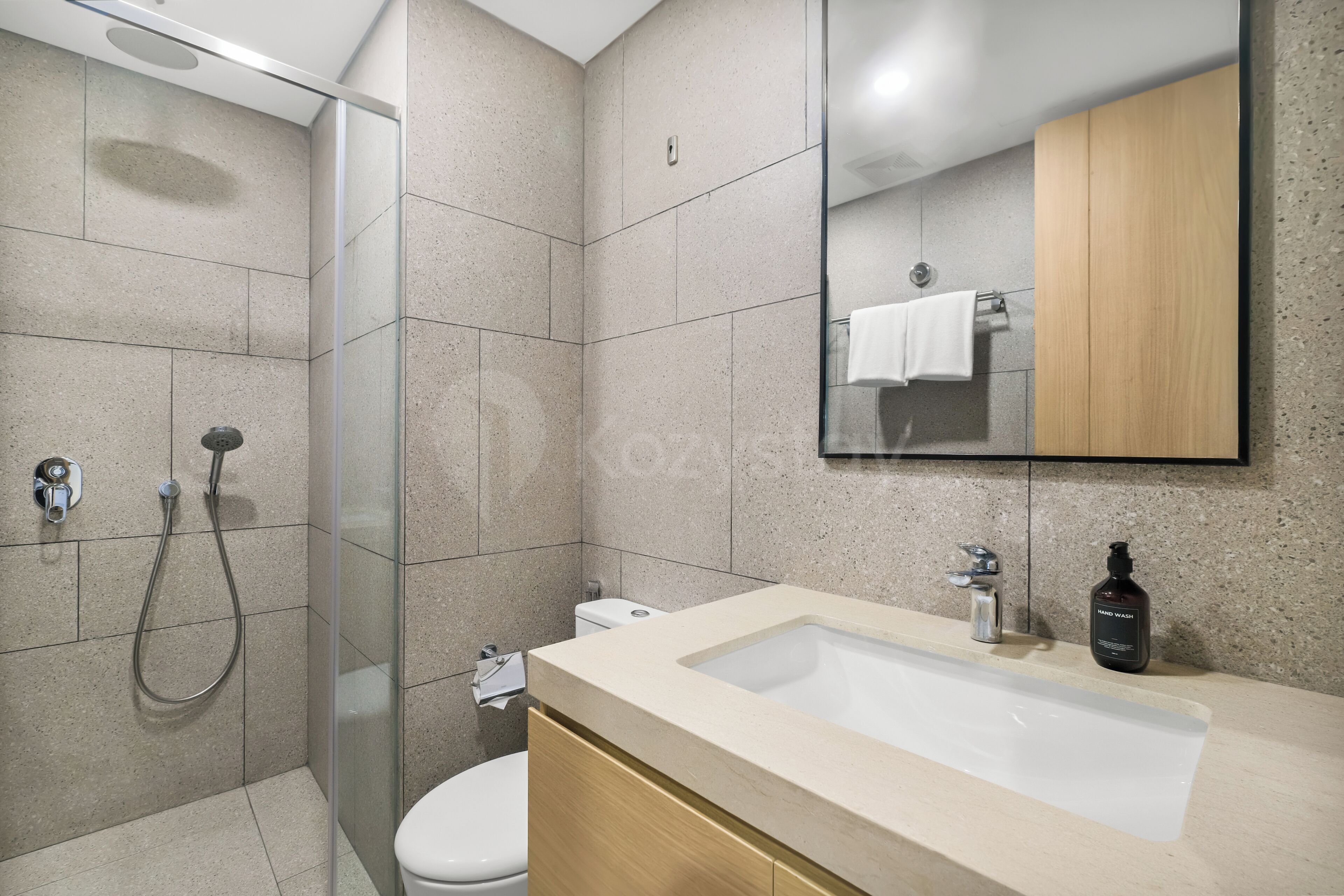A modern bathroom with a glass-enclosed shower, a white toilet, a beige sink with a mirror above, and a bottle of hand wash.