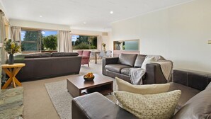 A modern living room with a sofa, armchairs, and a coffee table. There is a dining area with a table and chairs, and a large window providing a view of the outdoors.