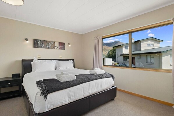 A neatly made bed with white linens and a gray throw, a nightstand with a radio, a painting on the wall, and a window with a view of houses and mountains. - Wanaka Lakeview | Games Room | Sleeps 8 (Wānaka)