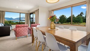 A dining room with a wooden table, white chairs, and a vase of flowers. There is a sofa and a window with a view of mountains and trees.