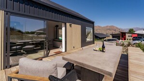 A modern outdoor living area with a wooden deck, a glass sliding door leading to a room, and a view of the mountains in the background.