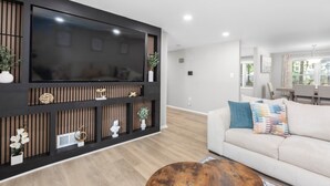 A modern living room with a large flat-screen TV mounted on a dark wooden wall, a beige sofa with colorful cushions, and a round wooden coffee table.