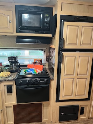 Cocina compacta con microondas, estufa y refrigerador negros, armarios de madera y una ventana con persianas.