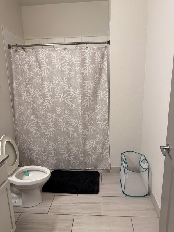 Un baño con inodoro, cortina de ducha estampada con hojas, alfombra de baño negra y cesto de ropa de malla.