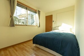 A bedroom with a bed, a window with curtains, a wooden floor, and a door.
