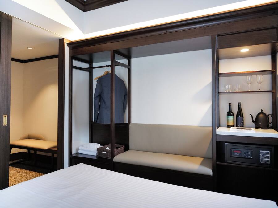 A hotel room with a bed, a seating area, a wardrobe, and a shelf with wine bottles and glasses.