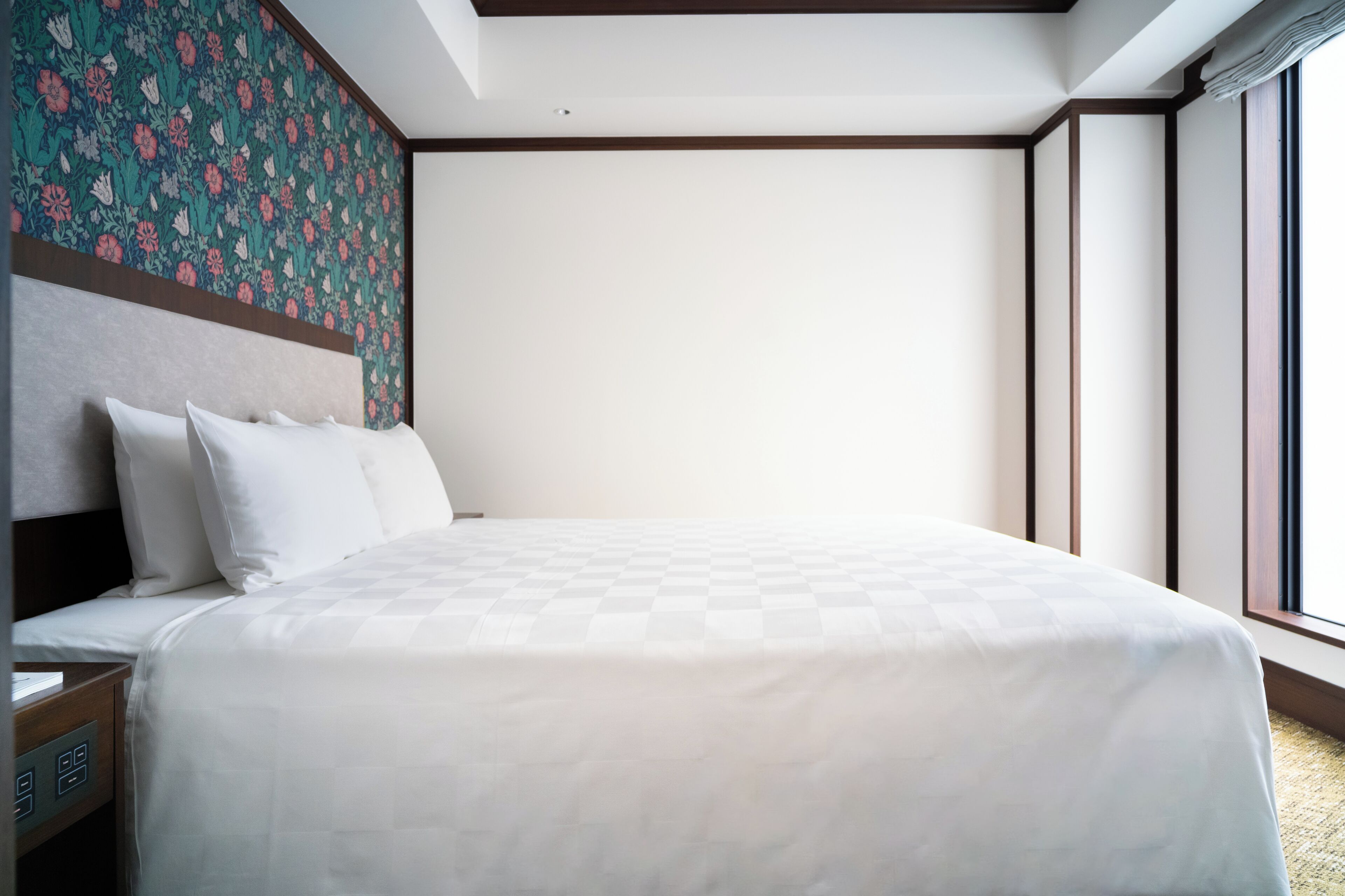 A neatly made bed with a checkered bedspread, a headboard with a floral pattern, and a nightstand with a power outlet.