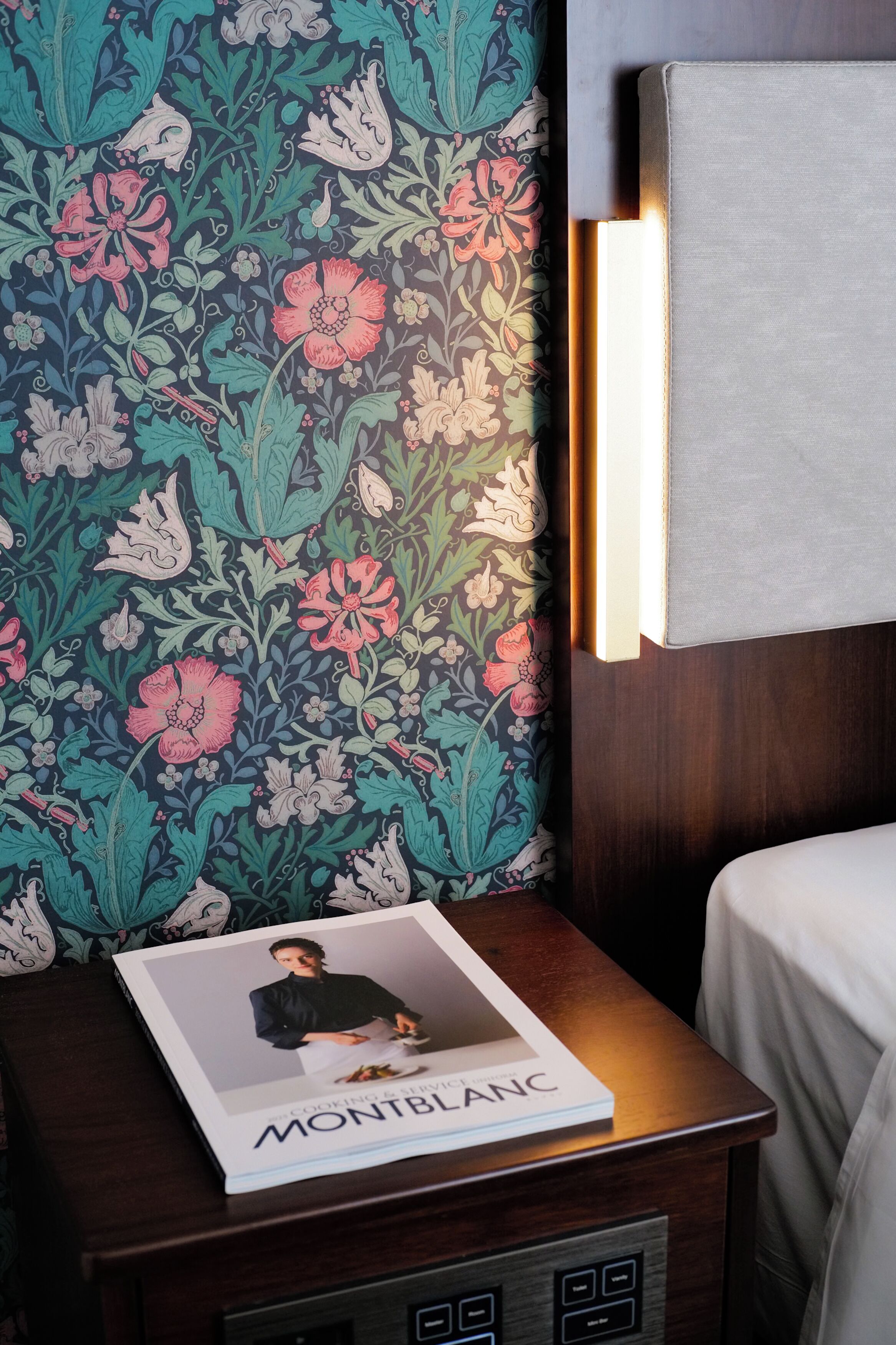 A neatly made bed with a white sheet, a bedside table with a book titled 'Montblanc', and a wall with a floral patterned wallpaper.