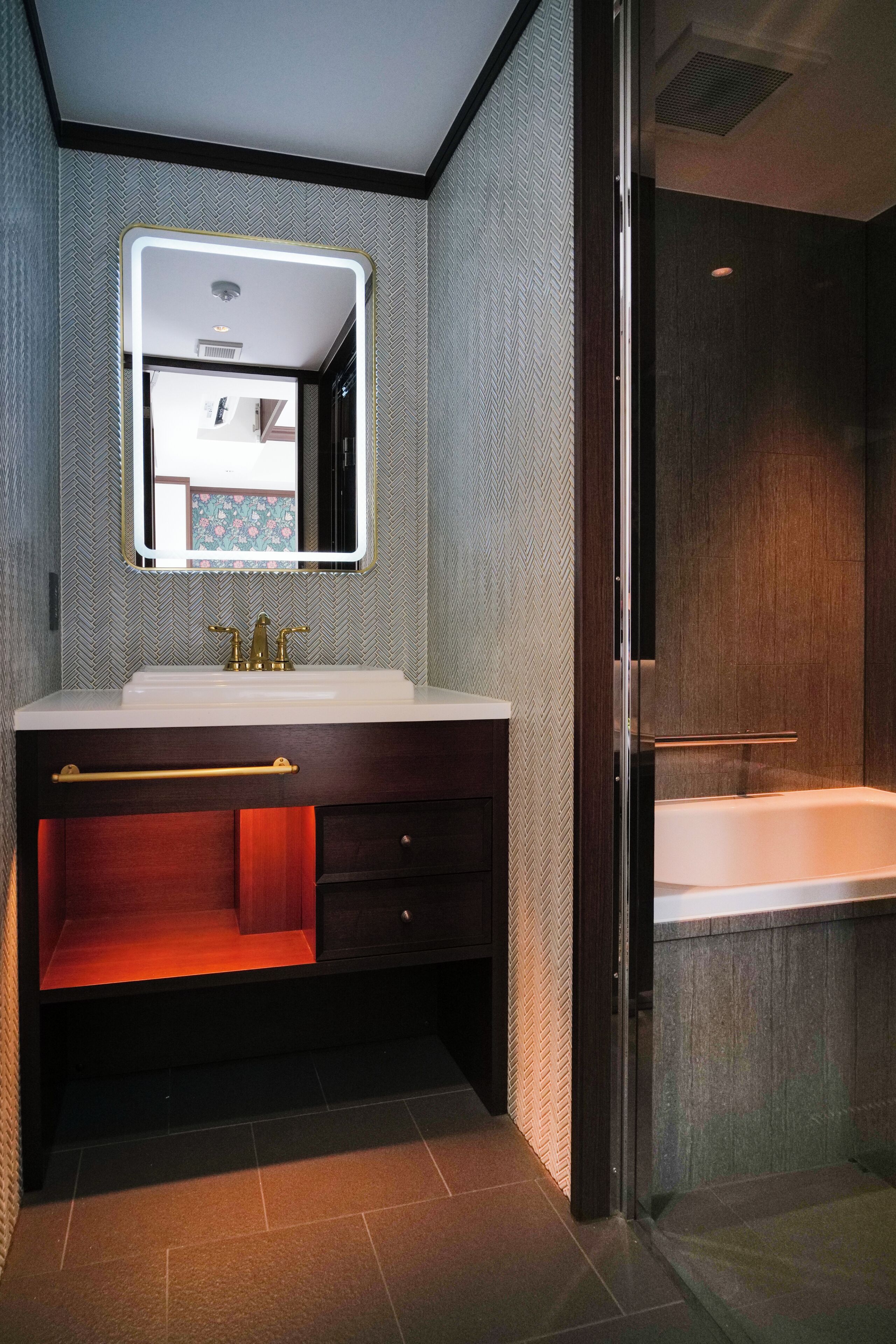 Un baño moderno con un espejo grande, un lavabo blanco, un mueble de tocador oscuro y una ducha con mampara de vidrio.