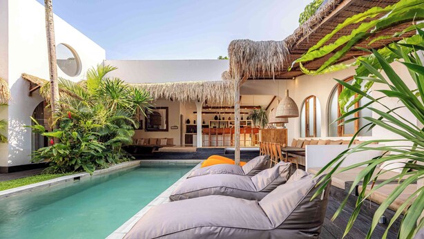 A pool area with lounge chairs, a thatched roof, and indoor seating.