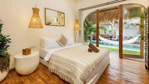 A bedroom with a large bed, a round side table, a painting, and a view of a pool and hammock.