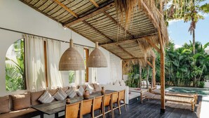 A spacious outdoor dining area with a long wooden table, cushioned seating, and hanging wicker lamps.