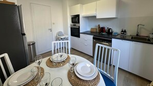 A compact kitchen with white cabinets, a microwave, an oven, and a dishwasher. There is a dining table set with plates, glasses, and a chair. - L'étape Aixoise - Terrace apartment car park Rotonde centre (Aix-en-Provence)