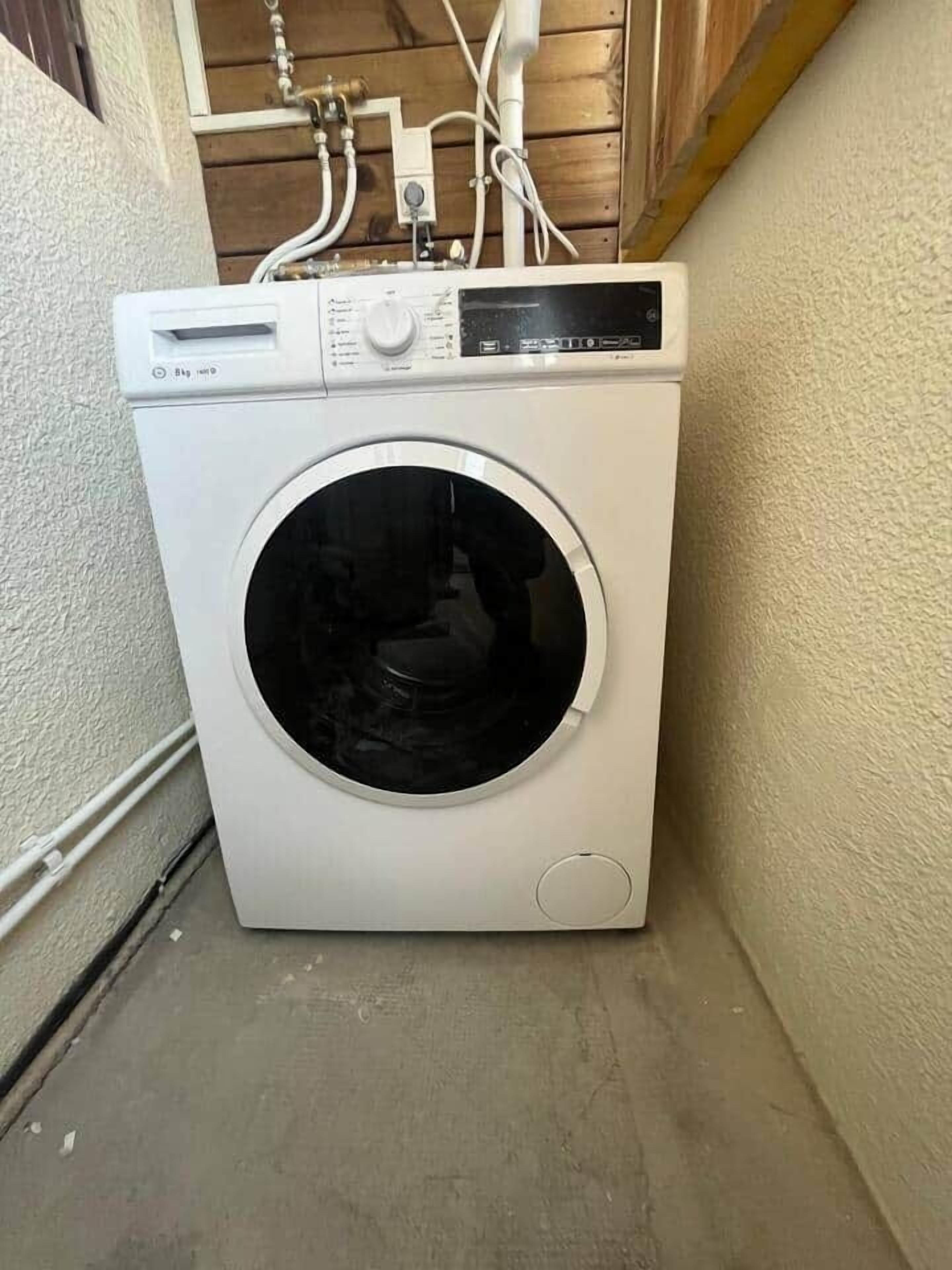 En tvättmaskin med frontmatning placerad i ett hörn, med en vägg av trä och en vit dörr.