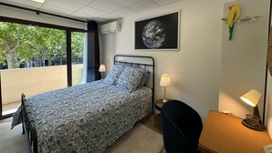 A bedroom with a bed, a desk, a chair, a lamp, and a window with a view of trees and buildings. - L'étape Aixoise - Terrace apartment car park Rotonde centre (Aix-en-Provence)