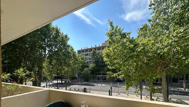 A balcony with two green chairs and a table set for two, overlooking a street with trees and buildings. - L'étape Aixoise - Terrace apartment car park Rotonde centre (Aix-en-Provence)