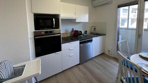 A compact kitchen with white cabinets, a microwave, oven, and dishwasher. There is a small dining table with chairs, and a window with a view of buildings. - L'étape Aixoise - Terrace apartment car park Rotonde centre (Aix-en-Provence)