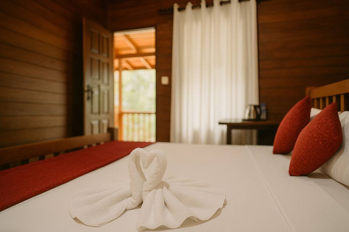 A neatly made bed with a heart-shaped towel, red pillows, and a wooden headboard.
