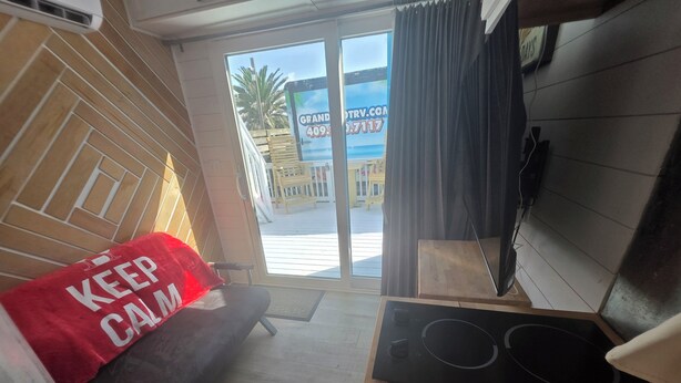 A room with a wooden floor, a black sofa with a red cushion, a small table with a black object, and a view of the outside through a glass door. - Coastal Caboose beach container home (Bolivar Peninsula)
