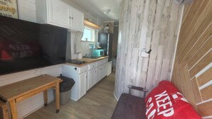 A compact kitchen with white cabinets, a microwave, and a sink. There's a wooden bench and a trash bin. - Coastal Caboose beach container home (Bolivar Peninsula)