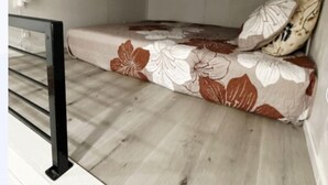 A small, compact room with a bed, a wooden floor, and a metal railing.