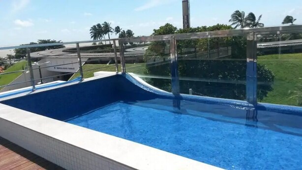 Una piscina con baranda de vidrio, rodeada de jardines con palmeras y vista a una carretera y edificios.