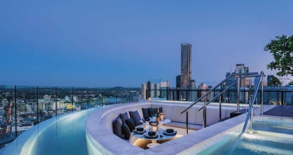 A rooftop infinity pool with a dining area, a city skyline in the background, and a glass barrier. - Valley Lookout | Luxe 1BR + Rooftop Pool + Parking (Fortitude Valley)