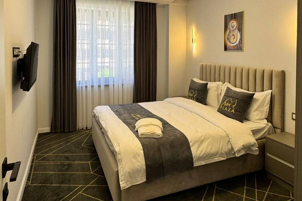 A hotel room with a bed, a flat-screen TV, an air conditioning unit, a window with curtains, and a geometric-patterned carpet.