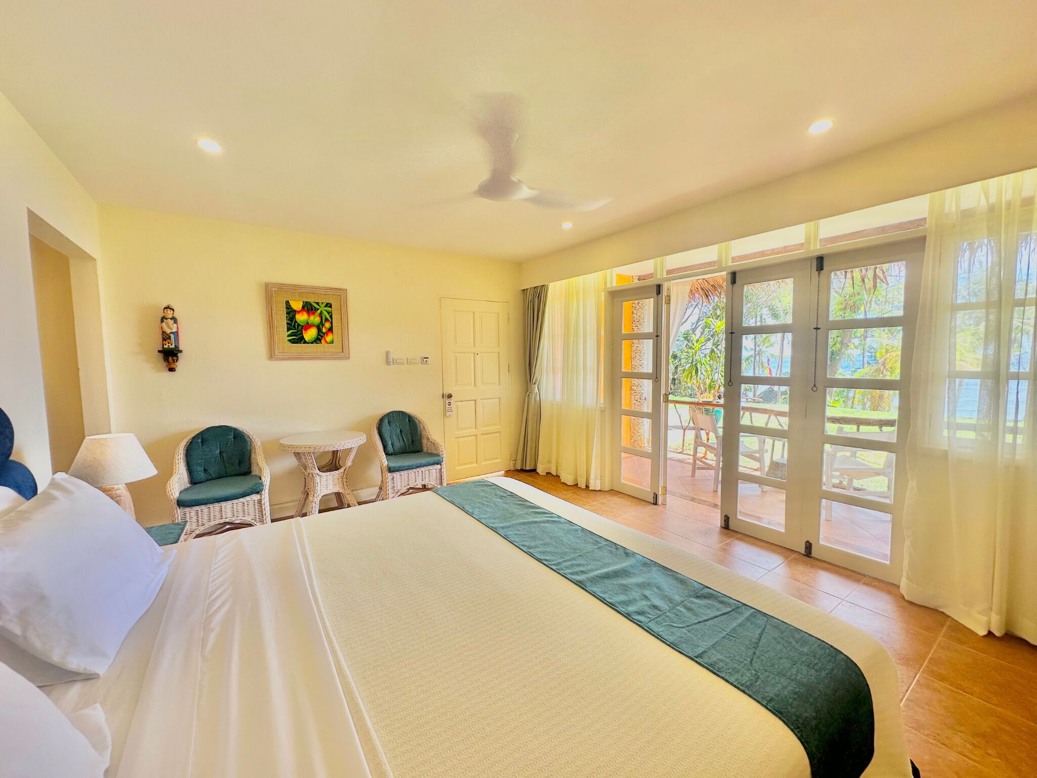 A hotel room with a bed, two chairs, a table, a ceiling fan, and a view of the outdoors through large sliding doors.