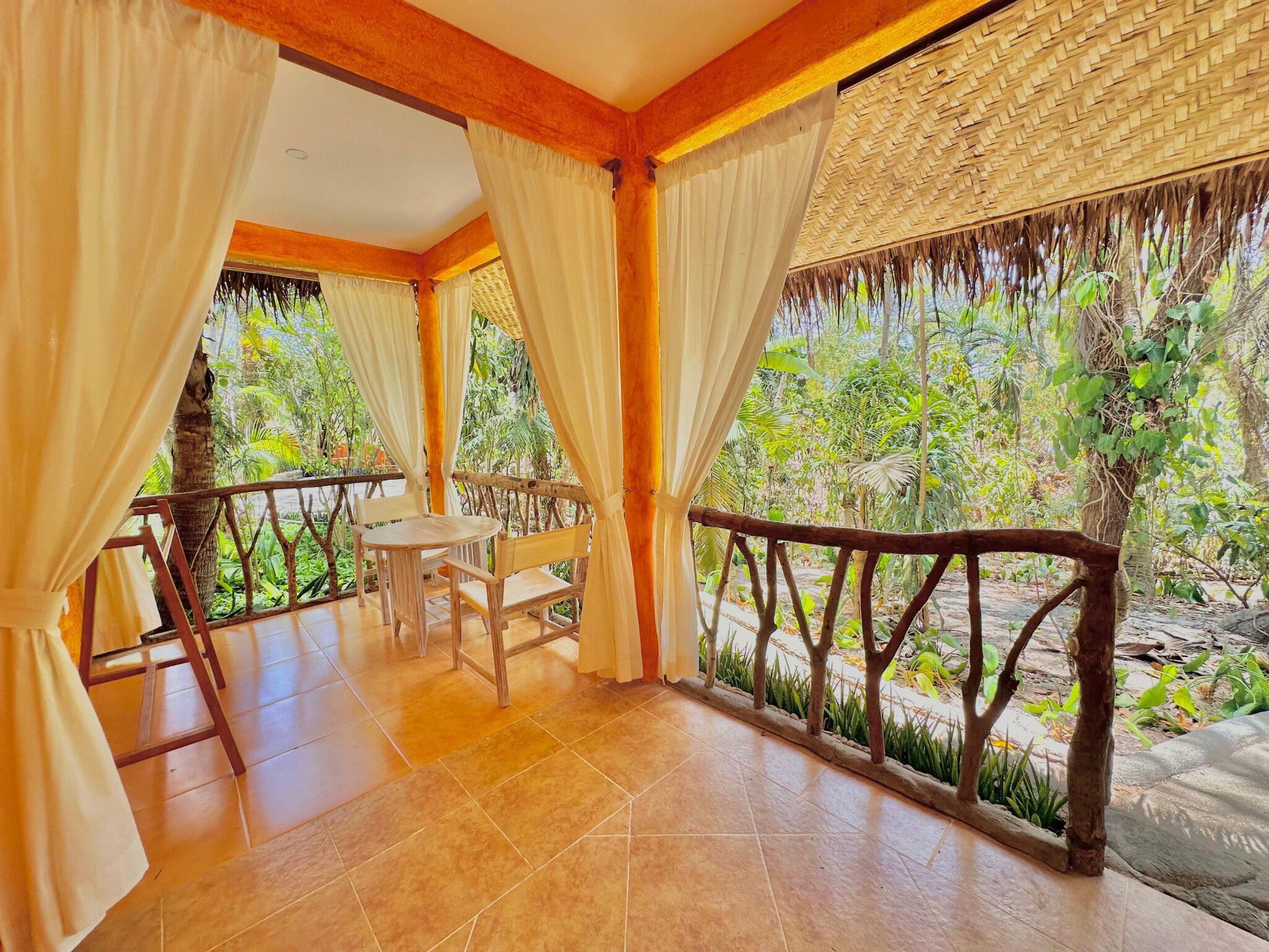 A room with a wooden table and chairs, a balcony with a railing, and a view of greenery outside.