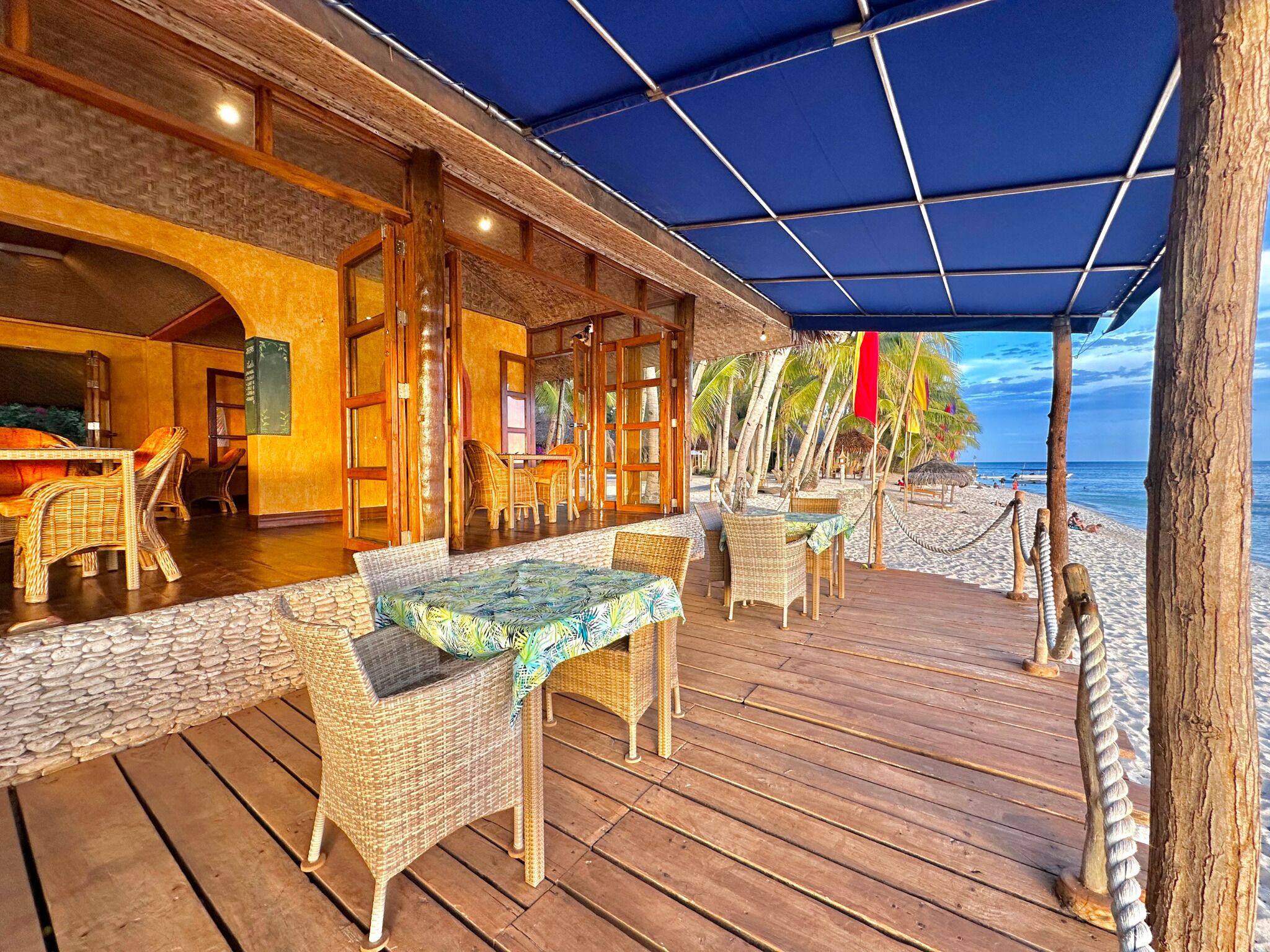 A wooden deck with wicker furniture, a covered area with a blue canopy, and a view of the beach and ocean.