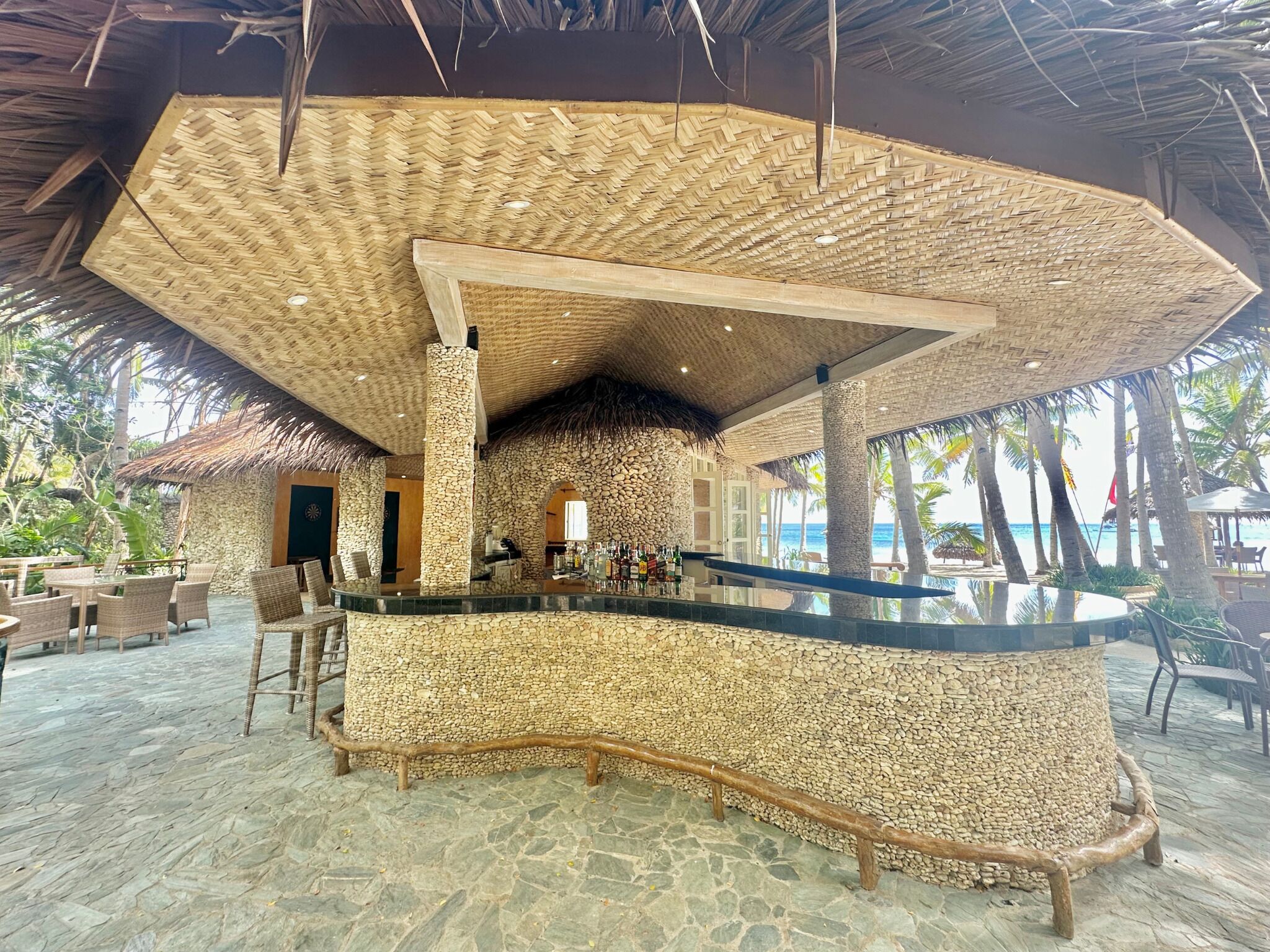 A beachside bar area with a stone wall, wooden furniture, and a view of the ocean.