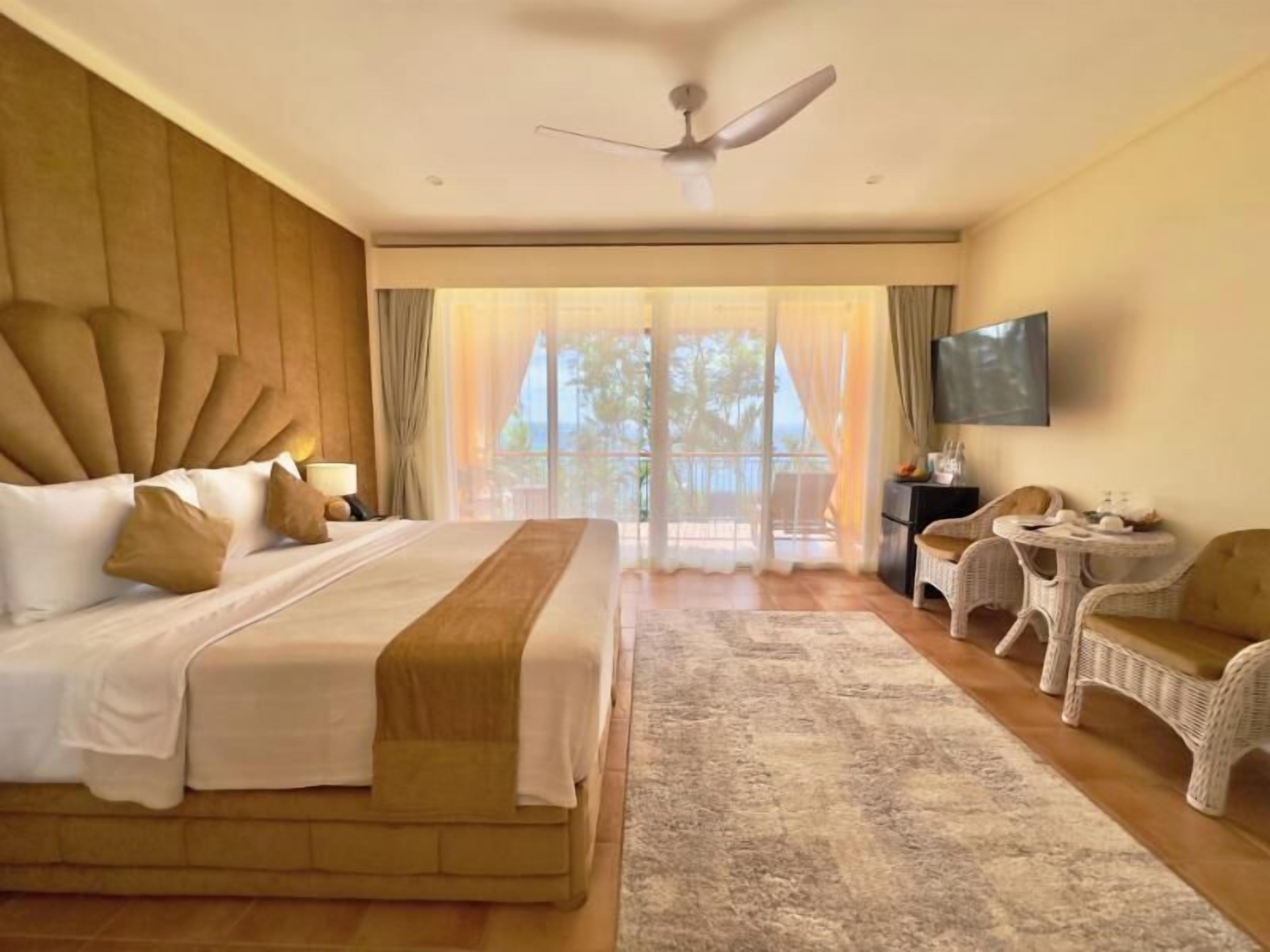 A hotel room with a large bed, a ceiling fan, a TV, wicker furniture, and a view through the window.