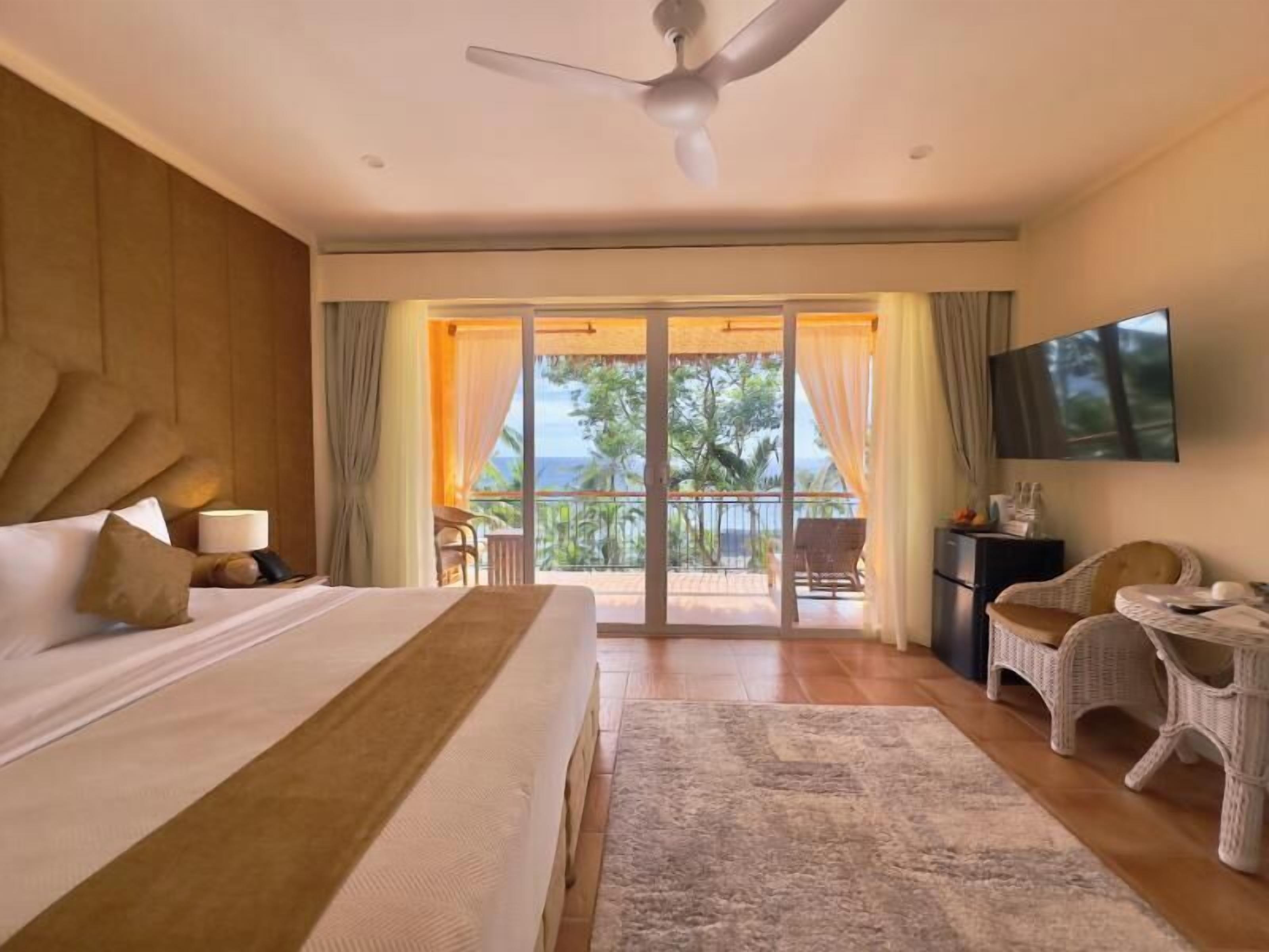 A hotel room with a large bed, a desk, a chair, and a balcony with a view of the sea.