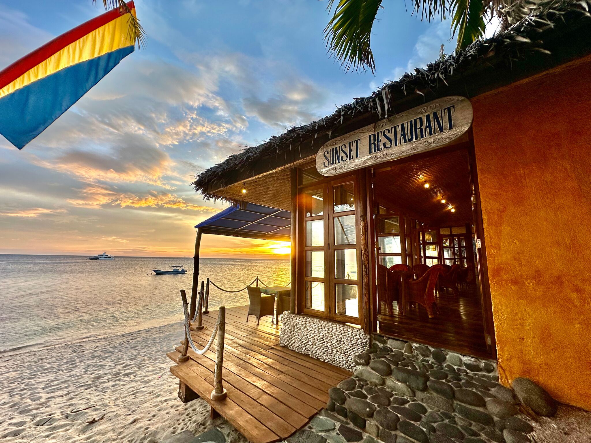 A beachside restaurant with a wooden deck, stone walls, and a sign reading "Sunset Restaurant."