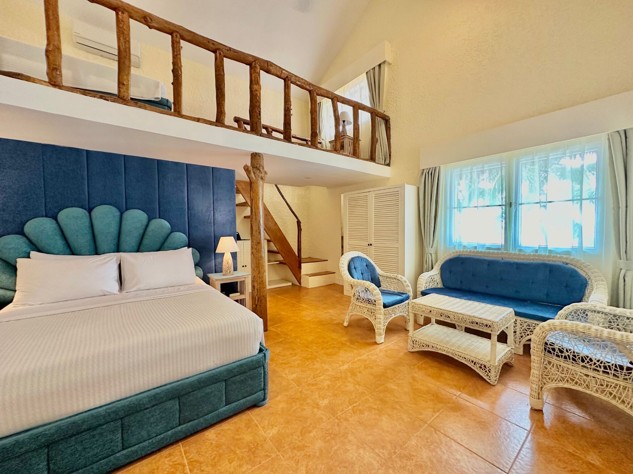 A bedroom with a bed, a blue upholstered headboard, a wooden staircase, a wicker sofa, and a window with curtains.