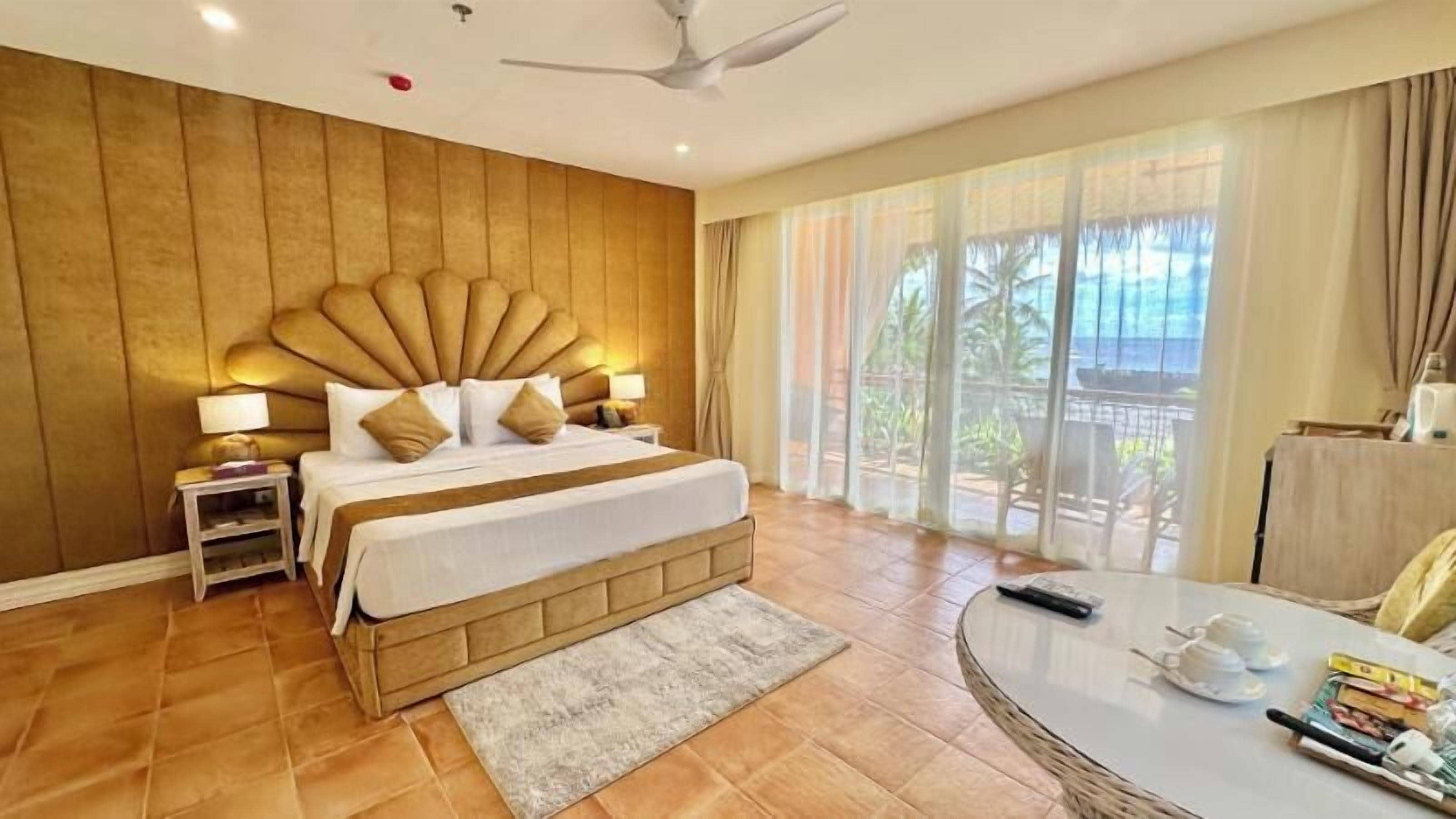 A hotel room with a large bed, a ceiling fan, a view of the outdoors, and a small table with items on it.
