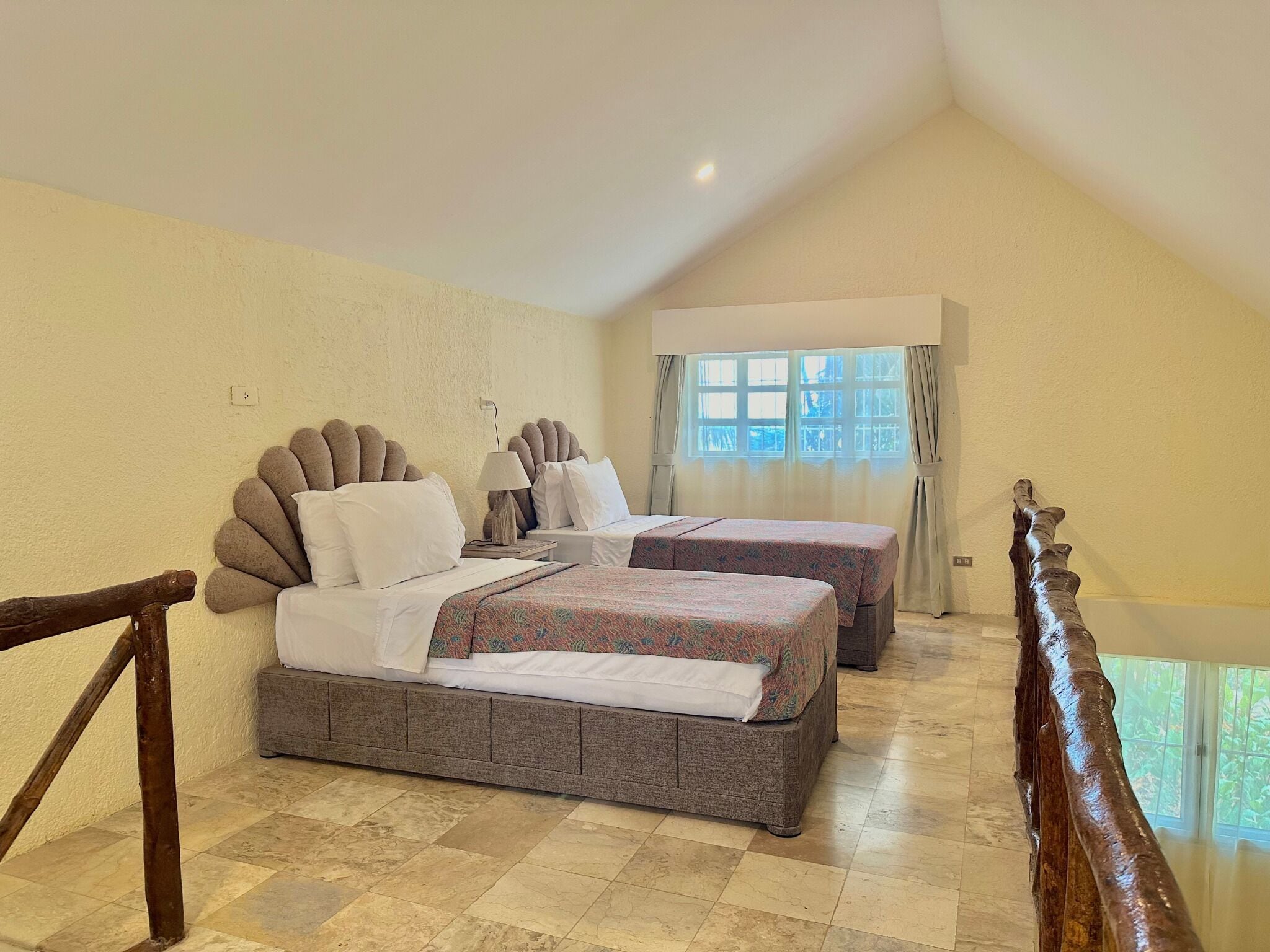 Quarto de hotel com duas camas, janela com cortinas, uma luminária e vista para o verde lá fora.