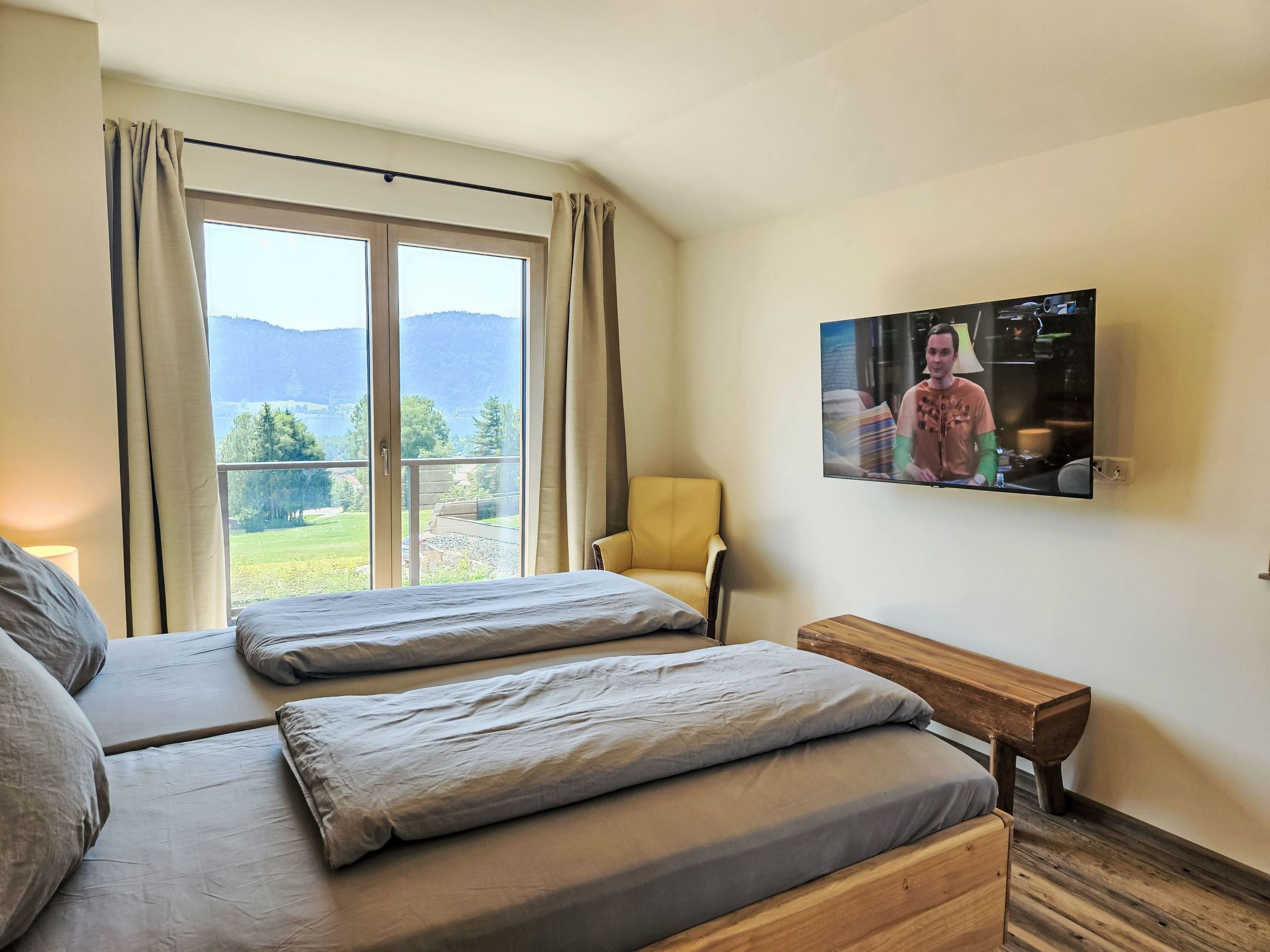 A hotel room with two beds, a wooden bench, a TV on the wall, and a large window with a view of mountains.
