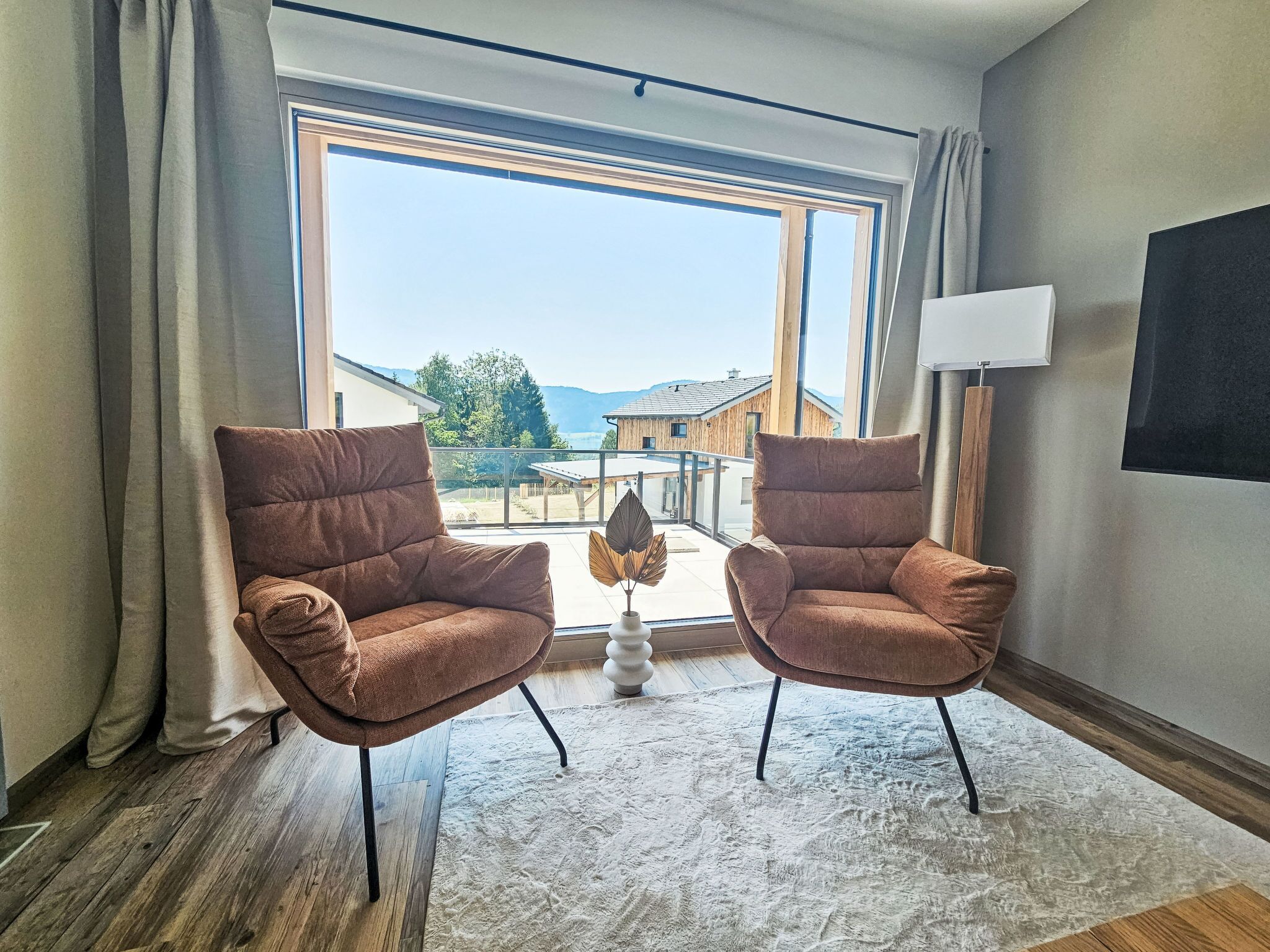 Une pièce avec deux fauteuils marron, un lampadaire blanc et une grande fenêtre donnant sur un balcon et des immeubles.