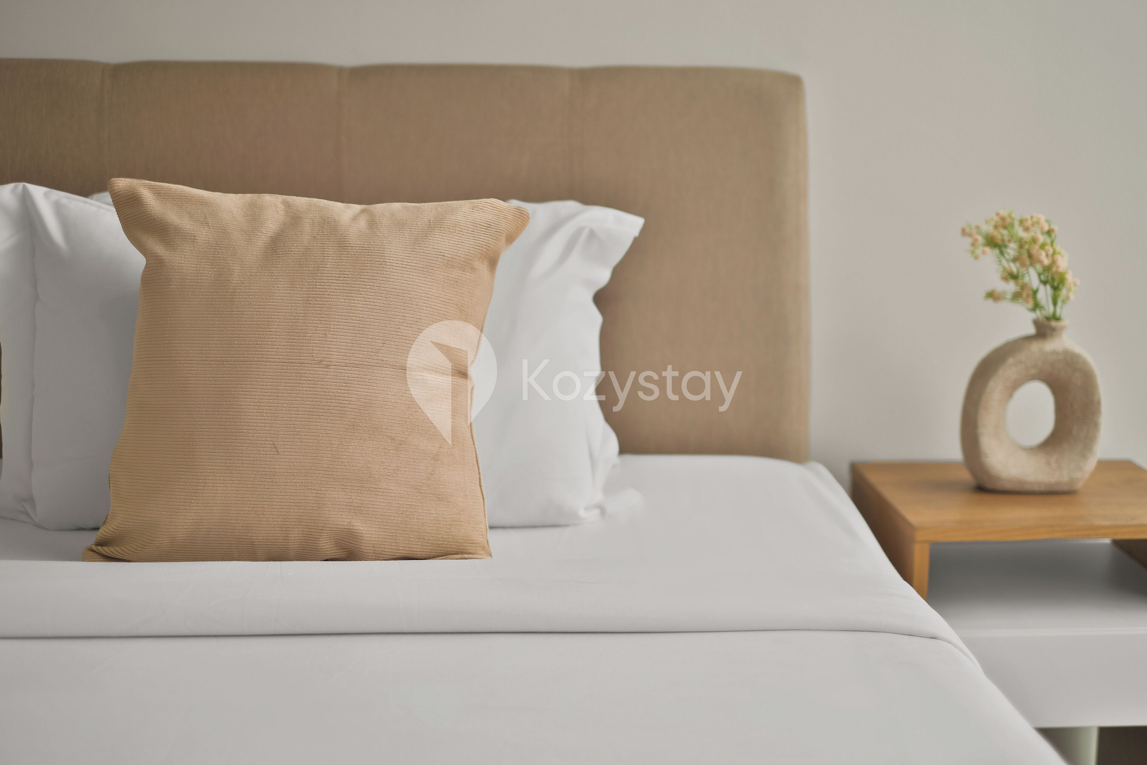 Ein Bett mit weißer Bettwäsche und einem braunen Kopfkissen, ein Nachttisch aus Holz mit Blumen in einer Vase und eine schlichte Wand im Hintergrund.