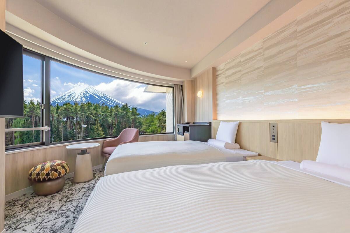 A hotel room with two beds, a desk, a chair, a TV, and a large window with a mountain view.