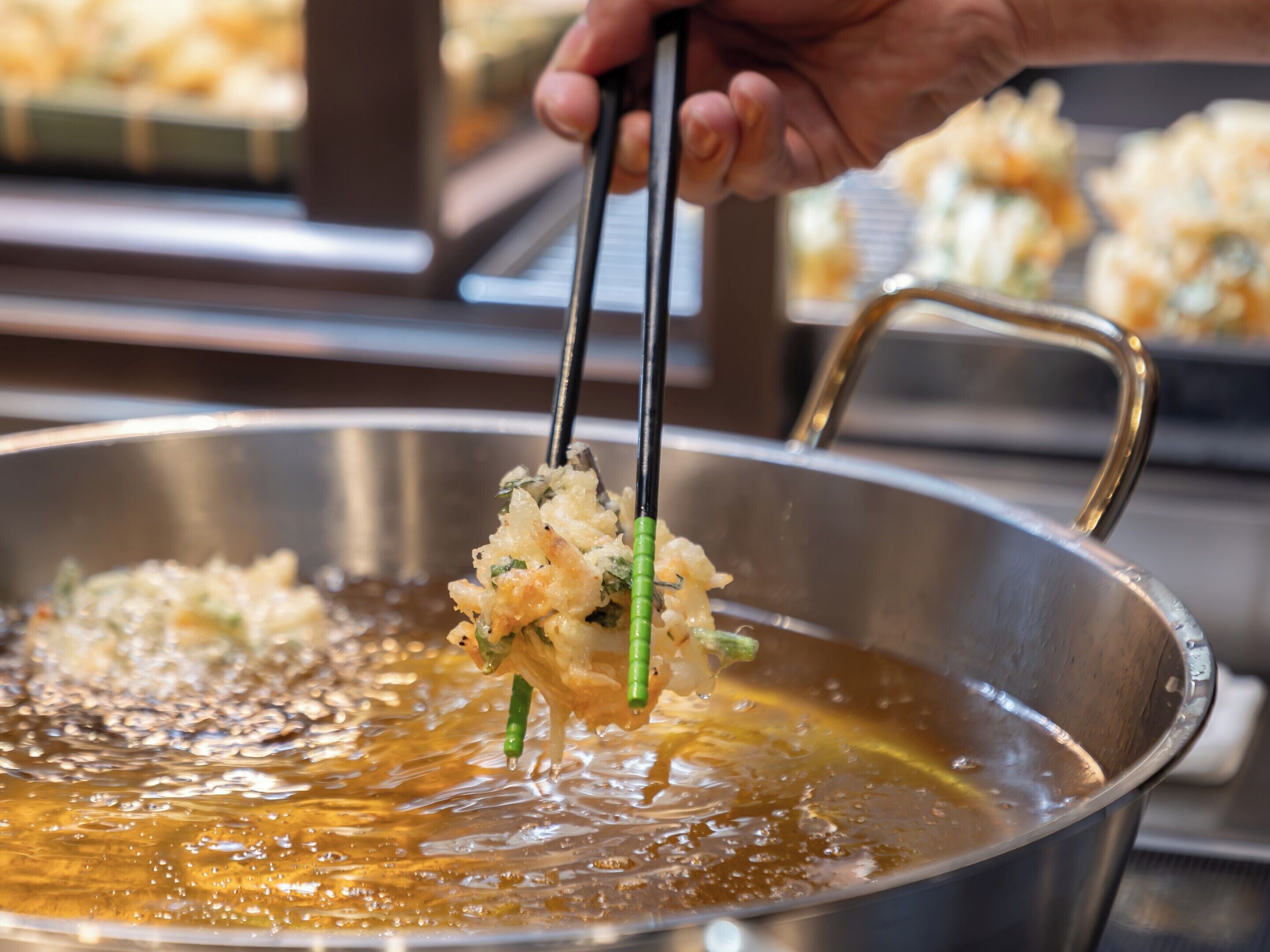 Una mano sosteniendo palillos sobre una olla con aceite y comida friéndose.